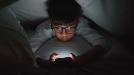 Close-Up-Of-Young-Boy-In-Bedroom-At-Home-Using-Mobile-Phone-To-Text-Message-Under-Covers-Or-Duvet-At-Night-2